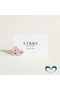 Tarjeta regalo LYKKE con flores naturales y texturas orgánicas en tonos beige y crema, mensaje "Disfruta de este momento. Gracias por ser parte de LYKKE" y logo de la marca
